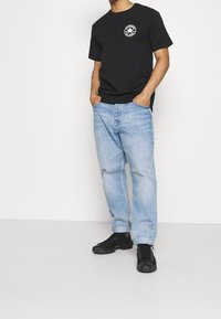 Osoba stojící s rukama v kapsách, oblečená v černém tričku s logem a světle modrých džínech, doplněná o černé boty, na neutrálním pozadí.