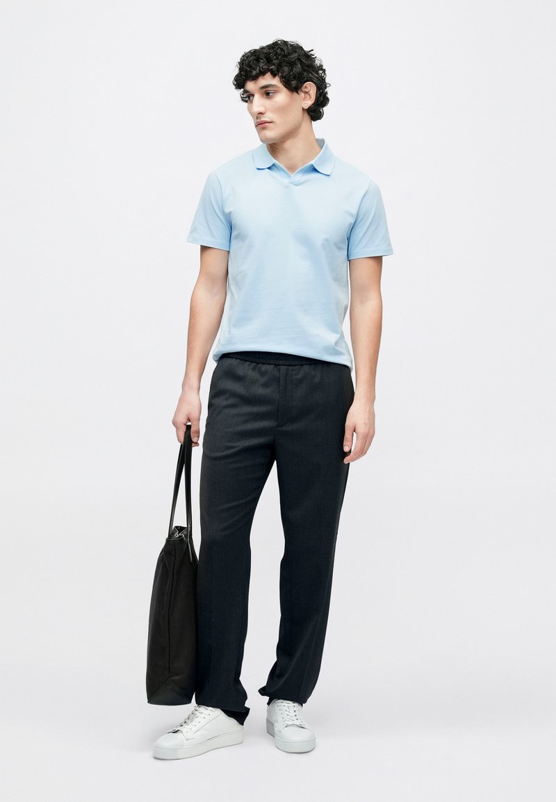 Hellblaues Poloshirt, dunkelgraue Hose und weiße Sneaker. Modell hält eine schwarze Tragetasche. Schlichtes Design, glatte Textur.