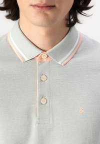 Polo gris clair avec un tissage texturé. Il présente un col rayé pêche, trois boutons, et un petit logo sur la poitrine.