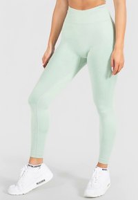 Muntgroene leggings met hoge taille, een gestructureerd patroon en een gladde afwerking, gecombineerd met witte sportieve sneakers met een logo.