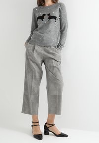 Femme portant un pull gris avec des motifs de chiens et des points blancs, un pantalon large court à carreaux, et des talons noirs à bride autour de la cheville, debout les mains dans les poches.