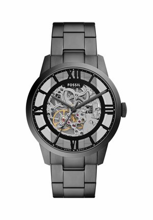 Montre-bracelet Fossil en métal noir avec cadran squelette, engrenages visibles, chiffres romains et trois aiguilles indiquant l'heure.