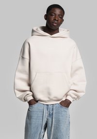 Lys beige hoodie med en frontlomme og broderet logo. Har dropped shoulders og oversized pasform, parret med lyseblå jeans.