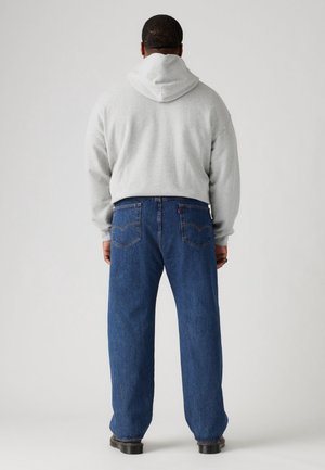 Mężczyzna zwrócony tyłem, ubrany w jasnoszarą bluzę z kapturem, luźne niebieskie dżinsy z tylnymi kieszeniami oraz czarne buty na jednolitym jasnym tle.