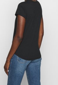 Person iført en ensfarvet sort kortærmet T-shirt og blå denim jeans, vist fra ryggen mod en hvid baggrund.