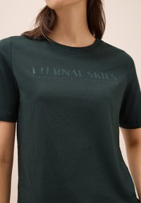 Camiseta de algodón en verde oscuro con cuello redondo, mangas cortas y texto en relieve "CIELOS ETERNOS REFLEJO DE PENSAMIENTOS" en el frente.