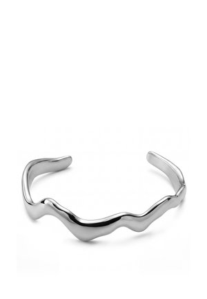 Hopeinen metallinen kädensijan rannekoru, jossa on sileä, epäsäännöllinen aaltomainen muotoilu ja avoimet päät, esiteltynä valkoisella pinnalla.