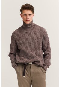 Brauner gerippter Rollkragenpullover aus strukturiertem Strickstoff; getragen mit beigen Hosen und einem Gürtel mit einer silbernen Schnalle.