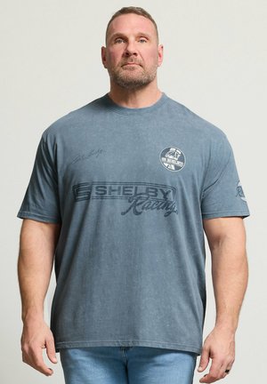 Homme aux cheveux courts et à la barbe portant un t-shirt Shelby Racing bleu délavé et un jean bleu clair, se tenant devant un fond uni.