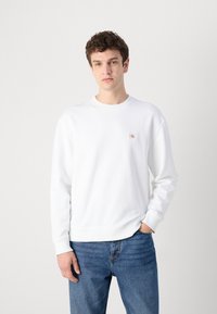 Sudadera blanca de algodón, con un cuello redondo y puños acanalados, con un pequeño logo de color en el lado izquierdo del pecho. Llevada con jeans azules.