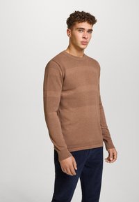 Maglione marrone lavorato a maglia con motivi testurizzati e maniche lunghe, caratterizzato da una scollatura a girocollo e polsini a coste, abbinato a pantaloni blu scuro.