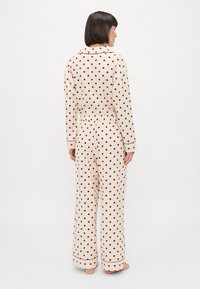 Cremefarvet pyjamasæt med røde prikker, der indeholder en skjorte med krave og bukser med elastisk talje. Blødt stof med piping-detaljer.