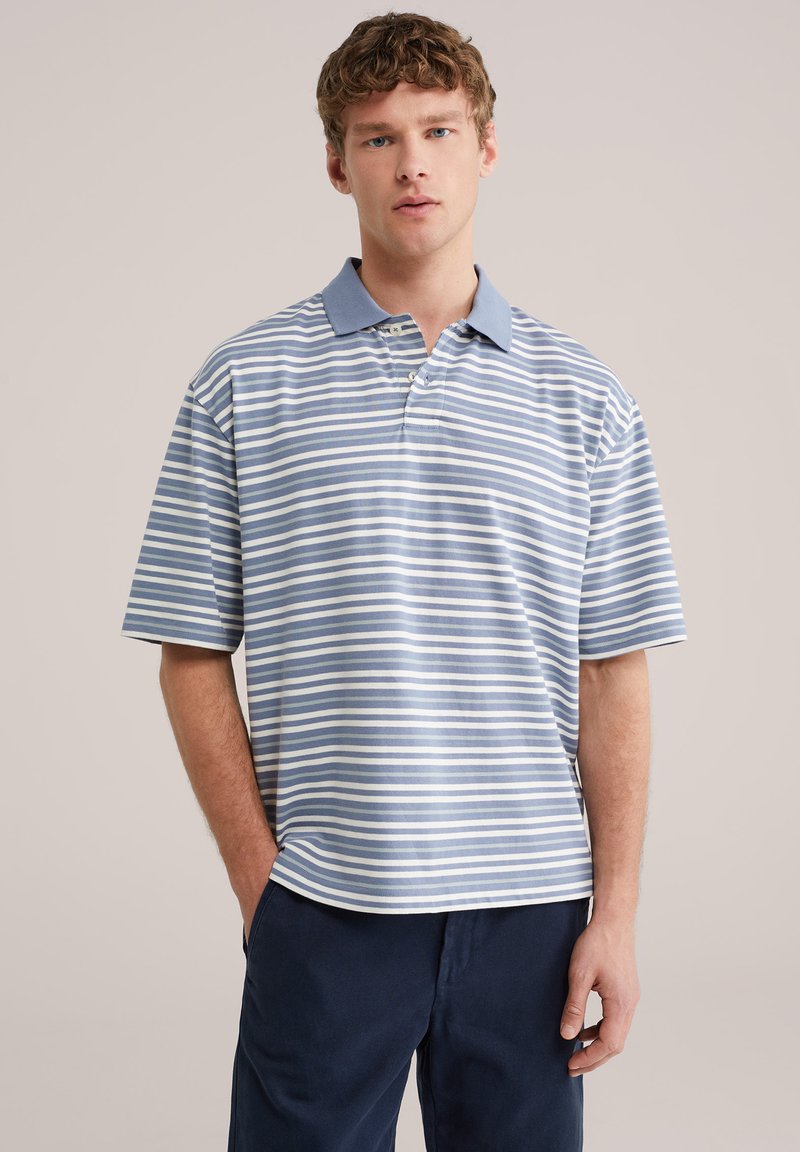 Jeune homme portant un polo à rayures bleu clair et blanches avec un col bleu et un pantalon bleu foncé, debout avec une main dans la poche.