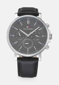 Montre en métal argenté, de forme ronde, avec un bracelet en cuir noir. Elle dispose d'un cadran gris foncé, de deux sous-cadrans, d'aiguilles argentées et d'indicateurs de date.