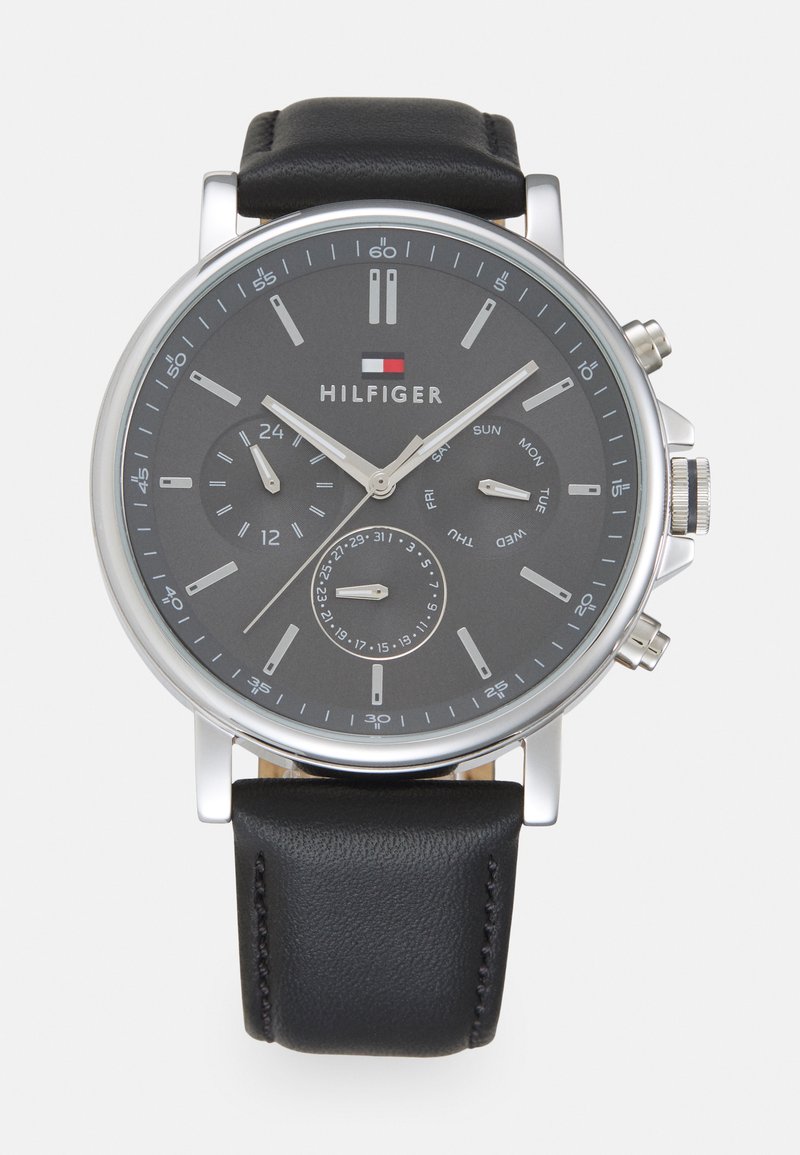 Montre en métal argenté, de forme ronde, avec un bracelet en cuir noir. Elle dispose d'un cadran gris foncé, de deux sous-cadrans, d'aiguilles argentées et d'indicateurs de date.