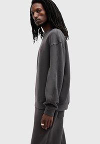 Sudadera gris con cuello redondo y mangas largas, que presenta una superficie texturizada y un sutil detalle de logo en burdeos. Combinada con pantalones grises.