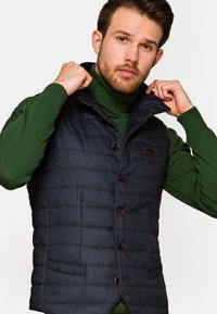 Granatowa kamizelka pikowana z zapięciem na guziki, płaskimi kieszeniami i wysokim kołnierzem, noszona na zielonym swetrze z golfem. Teksturowany materiał, dopasowany krój.