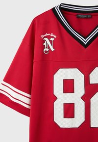 Maillot de sport rouge avec col en V rayé noir et blanc, numéro 82 blanc, bandes sur les manches et texte "Timeless Spirit" avec lettre N stylisée.