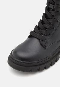 Bottine en cuir noir avec un bout rond, des lacets plats noirs, et une semelle en caoutchouc texturé avec des crampons prononcés.