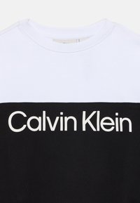 Sweatshirt Calvin Klein avec une section supérieure blanche et une section inférieure noire. Grand logo blanc sur le devant. Texture en coton lisse.