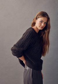 Mørk tekstureret strikket sweater med løs pasform og ribbede ærmegab, kombineret med skræddersyede grå bukser. Modellen har langt hår og en neutral baggrund.