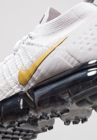 Witte Nike-sneaker met een textuur bovenkant, gouden accent aan de zijkant, en een transparante, volumineuze zool met duidelijke tractionpatronen.