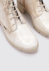 Botas de tobillo de color beige claro, hechas de cuero texturizado, con un diseño de cordones, tacones gruesos y una punta suave con un sutil brillo.