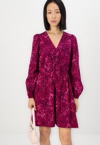Robe florale fuchsia avec de longues manches bouffantes, un décolleté en V profond et des détails à cordon. Modèle tenant un sac à main rose clair.