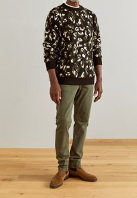 Pull en tricot vert olive foncé avec motif crème, associé à un pantalon vert olive clair et des bottines Chelsea en daim marron. Fond en parquet en bois.