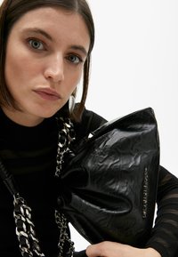 Eine Frau mit kurzen Haaren, die ein schwarzes Outfit und auffällige Ohrringe trägt, hält eine schwarze Tasche mit einer Kettenriemen und schaut intensiv in die Kamera.