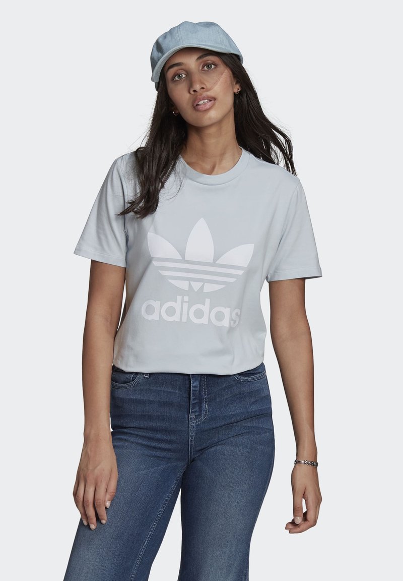 Šviesiai mėlynas medvilninis marškinėliai su dideliu baltu Adidas logotipu. Modelis juos derina su tamsiai mėlynomis džinsinėmis kelnėmis, demonstruodamas paprastą stilių.