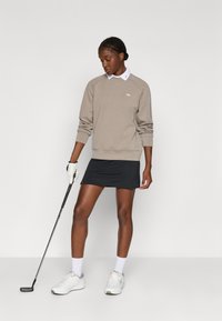 Beige színű pulóver kerek nyakkal, fekete golf szoknya, fehér cipők, és egy golf ütő. A pulóver bal felső részén logó található.