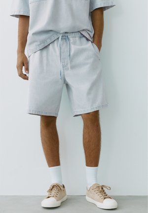 Homme portant un short en jean clair, une chemise ample assortie, des chaussettes blanches montantes et des baskets beiges, debout contre un mur blanc.