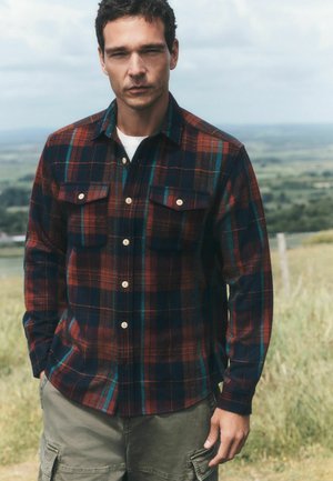 Man standing outdoors wearing a red and blue plaid button-up shirt and green cargo pants with a rural landscape in the background.