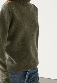 Close-up of a person wearing an olive green turtleneck sweater and blue jeans against a plain light background.