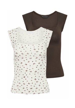 Donna con lunghi capelli scuri appoggiata al muro, che indossa un top marrone aderente senza maniche, jeans azzurri a gamba larga e scarpe bianche.