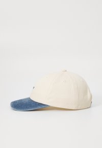 Baseball cap med et creamfarvet stofkrop og en blå denimskygge. Har luftventiler og et broderet logo på fronten.