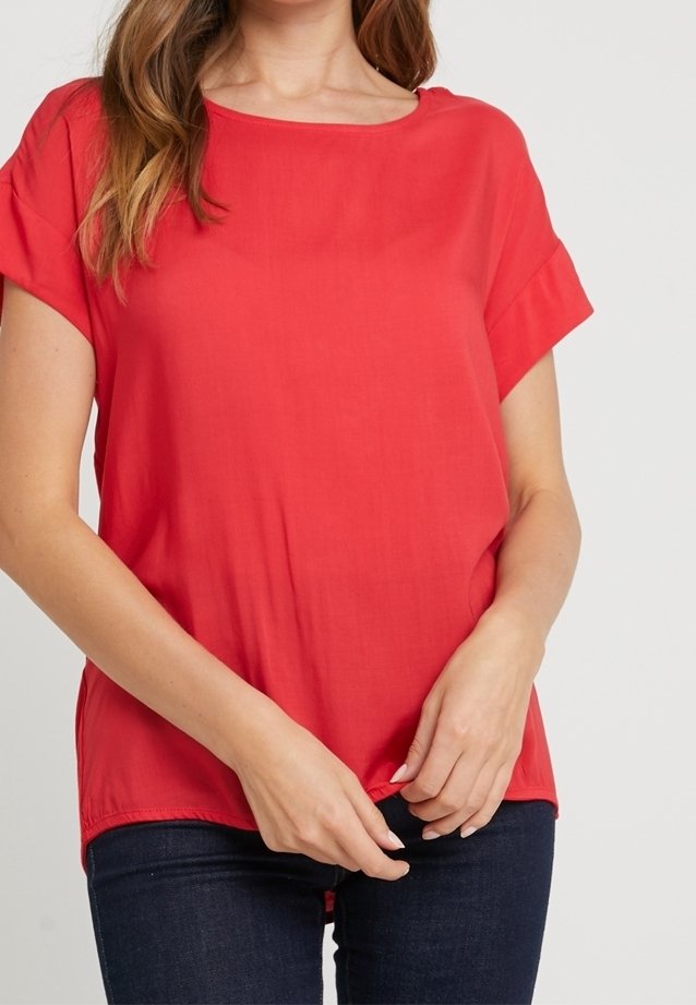 Femme portant un chemisier rouge vif à manches courtes et un jean bleu foncé, ajustant l'ourlet du chemisier avec les deux mains.