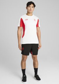 Biela atletická tričko s červenými akcentmi a logom Puma, spárované s čiernymi šortkami s červeným logom a detailmi po stranách.