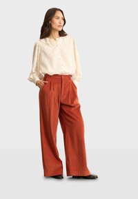 Blusa crema con colletto a volant e chiusura con bottoni, abbinata a pantaloni in corduroy ruggine a gamba larga con pieghe e tasche laterali. Scarpe nere.