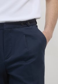 Homme portant un pantalon bleu marine avec des plis et deux boutons noirs à la taille, la main reposant dans la poche, associé à une chemise blanche texturée.