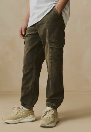 Pantaloni cargo din denim verde măslin cu manșete elastice, având multiple buzunare și o croială lejeră, asortați cu pantofi sport bej.
