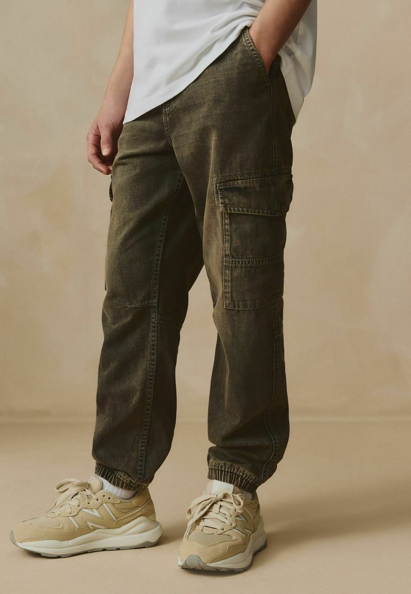 Pantaloni cargo in denim verde oliva con polsini elasticizzati, dotati di più tasche e una vestibilità rilassata, abbinati a scarpe da ginnastica beige.