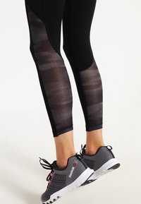 Czarne legginsy sportowe z siateczkowymi panelami, o fakturowanym wzorze. Noszone z szarymi butami sportowymi z różowymi akcentami i białymi podeszwami.
