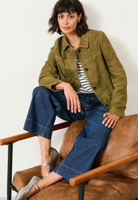 Woman sitting on a brown leather chair wearing an olive green suede jacket, striped shirt, wide-leg jeans, and silver flat shoes.