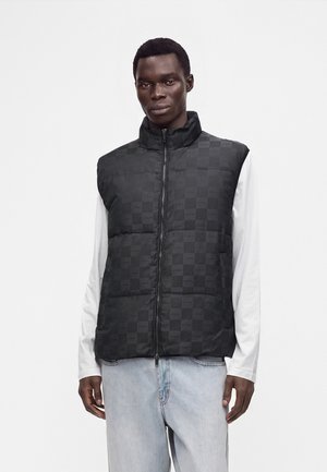Homme portant une veste matelassée noire à carreaux avec une fermeture éclair sur le devant, par-dessus une chemise blanche à manches longues et un jean bleu clair sur un fond uni.