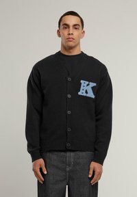 Svart cardigan med V-hals, fem knappar och en ljusblå "K"-patch med stjärnor på vänster bröst. Ribbad mudd och nederkant.