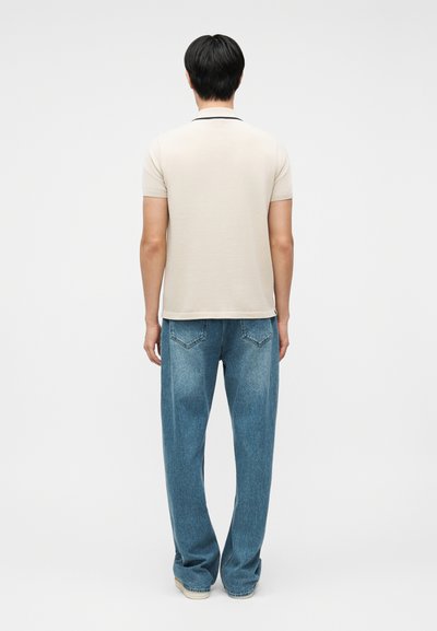 Άνδρας που έχει την πλάτη του προς το φακό, φοράει μπεζ πόλο με κοντά μανίκια και χαλαρά μπλε τζιν, μπροστά σε απλό λευκό φόντο.