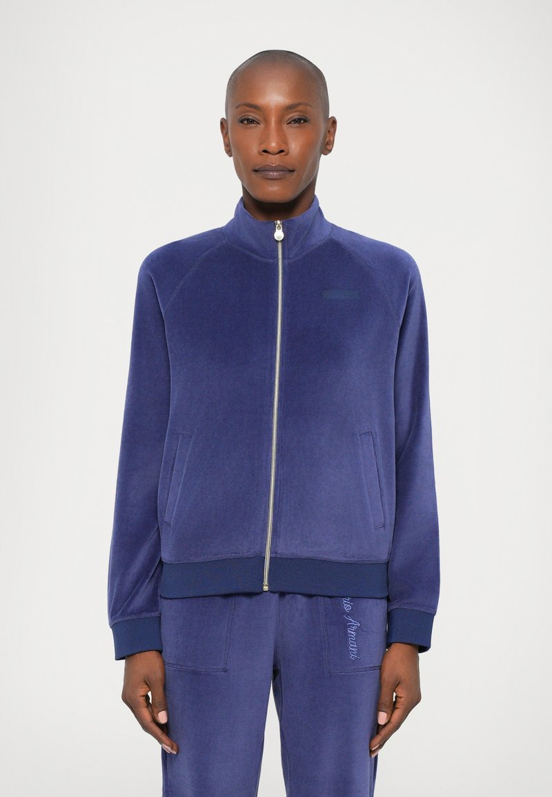 Chaqueta de cremallera en color navy, confeccionada en un suave tejido de terciopelo, con cuello alto, bolsillos laterales y puños y dobladillo acanalados. Presenta un sutil detalle del logo.