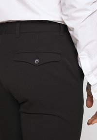 Svarta skräddarsydda byxor med en slät textur, figurnära design och en bakficka med knapp och rundad klaff.